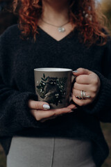 A girl in a warm sweater and red hair holds a cup of coffee that shows a raccoon