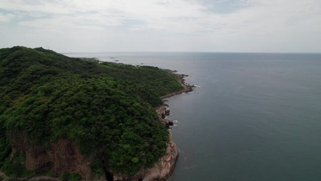 Drone orbits from left to right over Punta de Mita's rocky coast, revealing mountains and sea in a sweeping view.