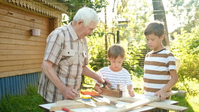 Elderly gray-haired senior man and two boys are standing at table with tools. Grandfather teaches his grandsons carpentry outdoors in the backyard of the house on a sunny day.