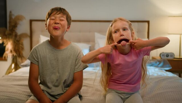 Funny happy Caucasian children kids boy girl brother sister siblings son daughter friends teasing camera at home grimacing playing together fooling around making faces showing tongue joking in bedroom