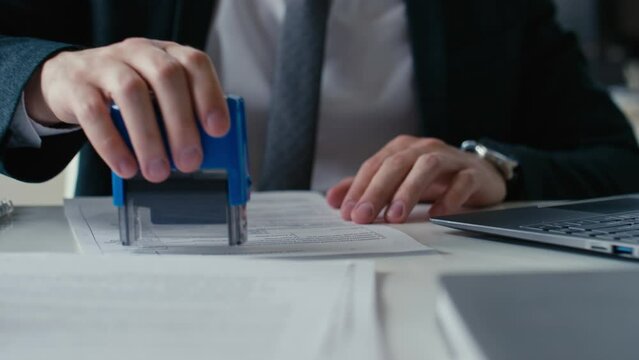 Closeup cropped stab shot of unrecognizable office worker approving documents with stamp and typing on laptop during workday in office