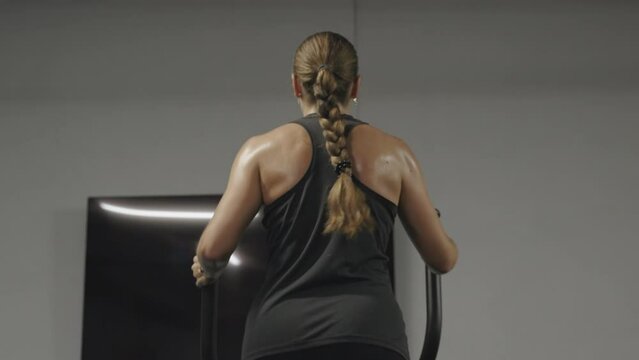 Sweat trickles down the back of a young woman working out in the gym on an Elliptical Trainer. Long hair braided. Weight loss, active lifestyle.