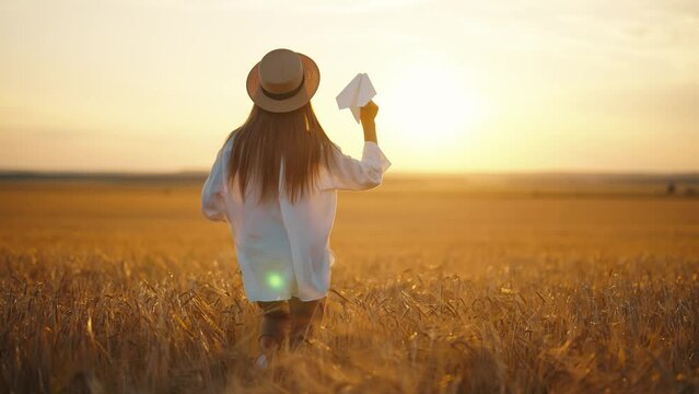 Carefree woman running on wheat field throwing paper plane in sky keeping straw hat on head at sunset, back view. Freedom, farm lifestyle, resting in countryside village, outdoor activities concept.