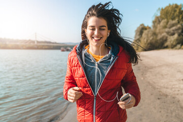 Young smiling woman wearing sports clothes and running on the beach while listening to music during...