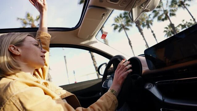 Blonde woman in car laughing and reaching out through sunroof, feeling of joy and freedom, sunny day with palm trees, concept of happiness. Singing favorite song and enjoying vacations driving on car