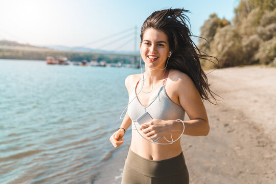 Young smiling sporty woman running on seaside while holding phone and listening to music. Slim fit female enjoying healthy morning routine on the beach.