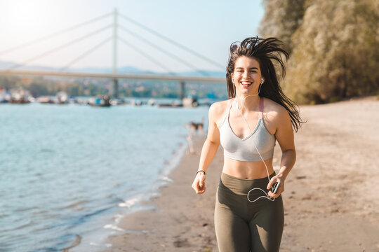 Young smiling fitness woman wearing sport bra and tights running on riverside while holding phone and listening to her favorite song or podcast.