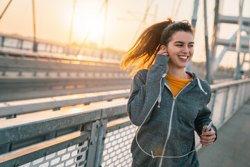Happy sportswoman listening to music on earphones while jogging at the bridge at sunrise during her...