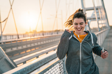 Obraz premium Dedicated smiling young woman with earphones running during her morning fitness routine and enjoying the music in urban environment.