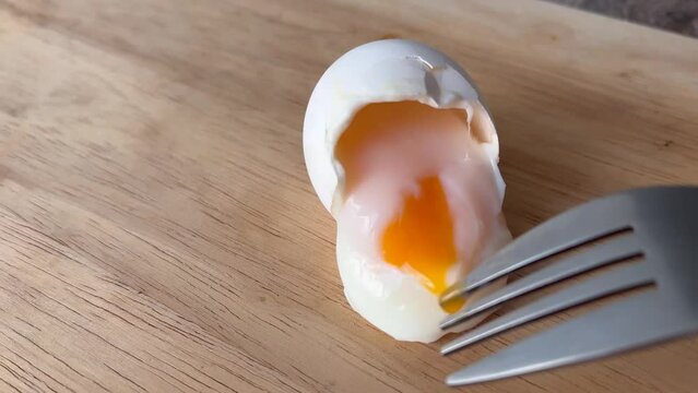 Liquid bright yellow yolk flows from boiled egg onto wooden surface of cutting board. Fork cuts egg white. Soft-boiled egg. Concept of healthy food, cooking breakfast, lunch, brunch, dinner, diet