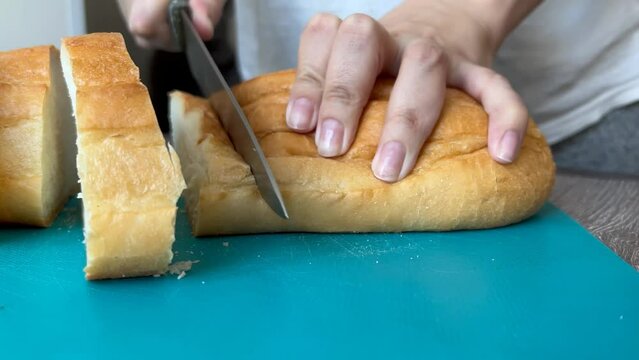 Woman cuts white bread with dull knife into large pieces for sandwiches on blue plastic cutting board. Girl cuts loaf poorly on wooden table in bright kitchen. Concept of preparing breakfast, lunch