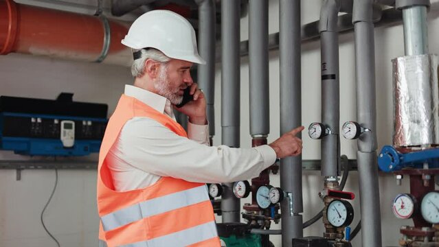 Side view of competent inspector pointing with finger at pressure indicators in pipes while talking on modern smartphone indoors. Mature gray bearded male working skillfully in protective clothing.