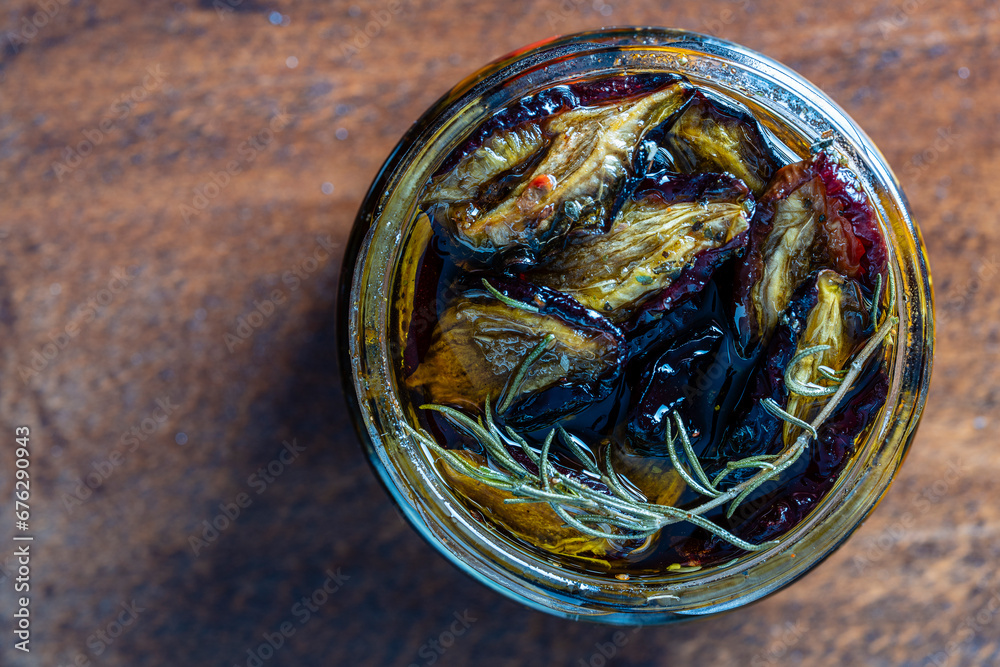 Wall mural Sun-dried plums with garlic, green rosemary, red chili pepper, olive oil and spices in a glass jar on a wooden table. Rustic style, top view, close up