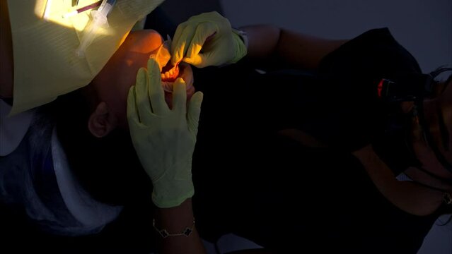 Slow motion vertical shot of a dentist wearing green latex applying resin glue on top of a patient's incisor tooth before placing a veneer brace. Yellow light