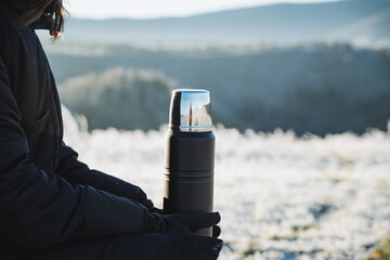 Black metal thermos on the background of winter forest, beautiful landscape, object in human hands, thermo flask for hot drinks. - obrazy, fototapety, plakaty