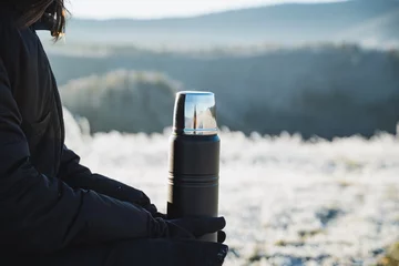 Fototapeten Black metal thermos on the background of winter forest, beautiful landscape, object in human hands, thermo flask for hot drinks. © Aleksey
