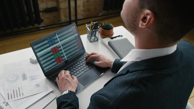 High angle over shoulder shot of businessman in suit stretching neck during sedentary office work on laptop with documents
