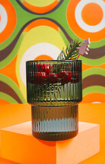 Emerald Green Ribbed Glass Cranberry Cocktail Drink with Pine Sprig on a Groovy Orange and Green...