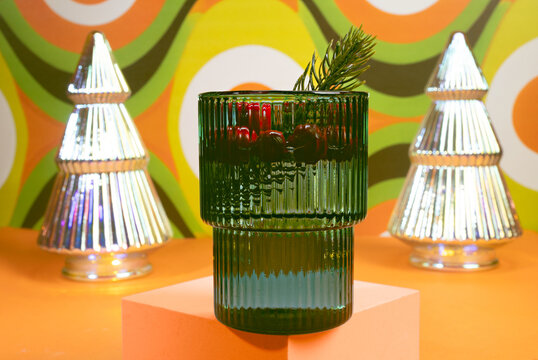 Holiday Shine: Emerald Green Ribbed Glass Cocktail Drink with Pine Sprig and Silver Metallic Christmas Trees on a Groovy Orange and Green Background