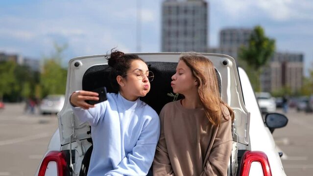 Beautiful happy teen girl sitting in car trunk with pretty mom taking selfie photos on smartphone. Real time video. Caucasian woman with daughter or sister making funny face expression posing to