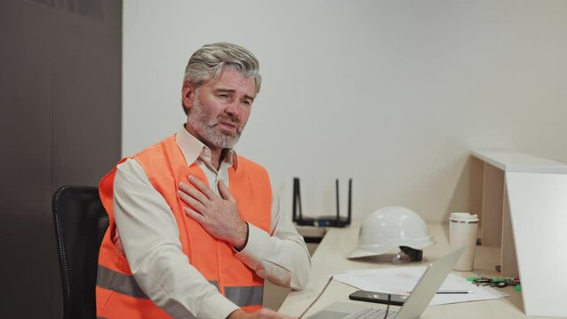 Aged caucasian man suffering from heart disease while sitting at office desk with laptop and smartphone. Exhausted male plant worker in orange vest closing eyes because of severe pain in chest.