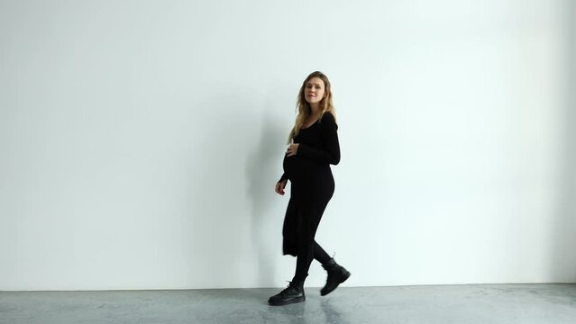 a pregnant girl in a black dress on a white background walks along a large wall.