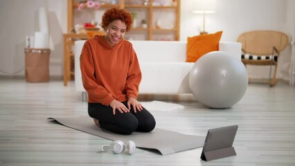 Smiling african american slim sporty girl warming wrists before online yoga practice sitting on mat at home looking at tablet screen. Redhead woman enjoying exercise. Online training, fitness concept.
