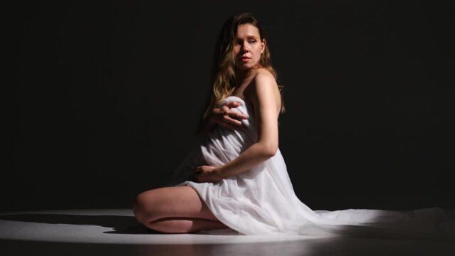 a beautiful pregnant girl dressed in a veil of white fabric sits on the floor on a black background