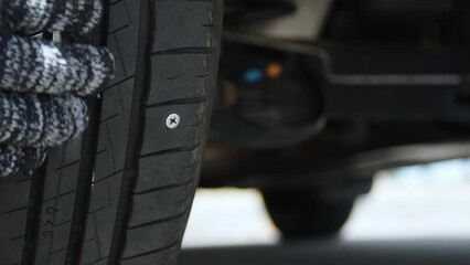 Driver, wearing protective gloves, rotates rear wheel and touches screw that punctured tire. Close up of damaged car tire with summer tread due to unsafe debris on road.