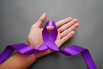 A hand holding a luxury purple ribbon on a gray background. Purple ribbon with pancreatic cancer...