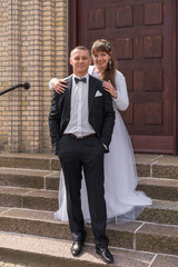 The bride and groom are standing on the steps against the backdrop of brown doors. The groom keeps his hands in his pockets. The bride stands behind the groom and smiles cheerfully. Weddin