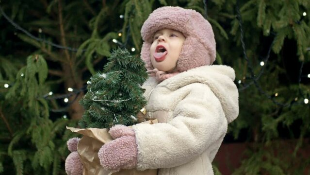 A 2-3 year old girl at the Christmas market holding a small Christmas tree. A child catches the falling snowflakes with her tongue.