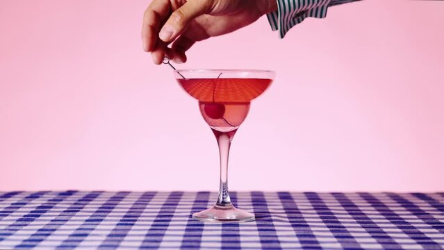 Glass with sweet Manhattan cocktail on checkered tablecloth over pink background. Putting brandied cherry for decoration. Concept of cocktails, alcohol drink, bar, taste, menu, pop art style.