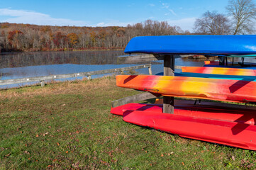Racks of colorful kayaks and canoes by state park autumn lake with woodland background and copy...