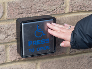 A brick wall mounted square pad is being pushed by a hand.Entry or exit point for disabled wheelchair user. Symbol and wording saying ,Press to Open'
