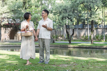Fototapeta premium Senior man and wife holding a picnic basket walking among the green trees in the park A retired couple in their 60s walked through the garden together enjoying their vacation.