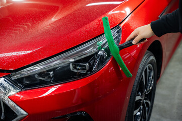 A man washes the headlights of a red car with a scraper. 