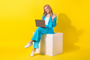 Full length photo of pleasant woman wear blue stylish suit sit on white cube talk on laptop video call isolated on yellow background