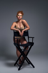 redhead plus size model in beige underwear standing near chair on dark grey backdrop, full length