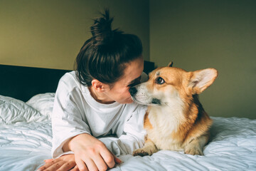 Girl lies on a bed with corgi dog at home. Welsh Corgi Pembroke kissing owner