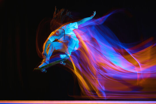 Young man, skateboarder with modern hairstyle practicing Ollie on skateboard with neon flashes of light isolated black background.