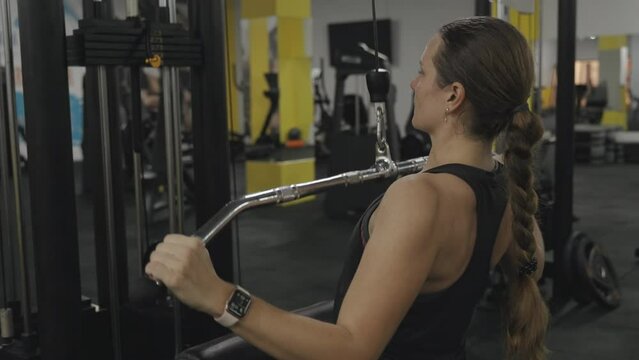 Young woman performs wide-grip seated lat pulldowns, developing the latissimus dorsi muscles and the shoulder girdle.