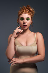 portrait of young redhead and curvy woman in bra and corset looking at camera on dark grey backdrop