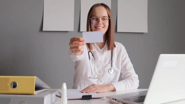 Professional woman doctor with stethoscope wearing white lab coat sitting in her office at table with laptop showing calling card with are for information while being at her workplace in stuff room.
