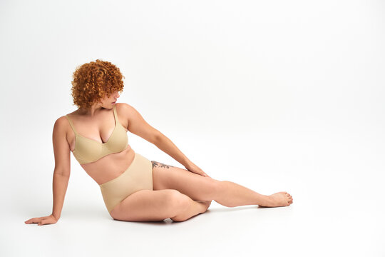 full length of redhead woman with curvy body sitting in beige underwear on white, plus size beauty
