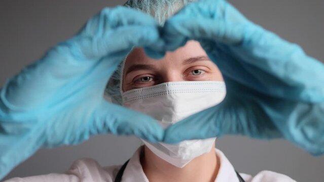 Smiling satisfied female doctor cardiologist or nurse wearing uniform medical cap gloves and face mask making heart with hands showing love gesture posing in hospital in stuff room.