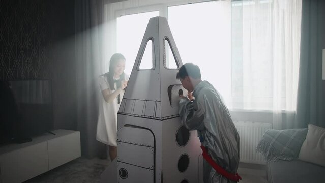 An Asian woman with son play in the living room at home, a boy in an astronaut costume with her mother, children together with their mother playing with a cardboard model of a spaceship.