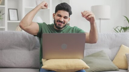 Hopeful hindu guy in casual attire crossing fingers in luck while expecting news from work. Bearded male sitting on couch, reading notification about promotion and making winner gestures.