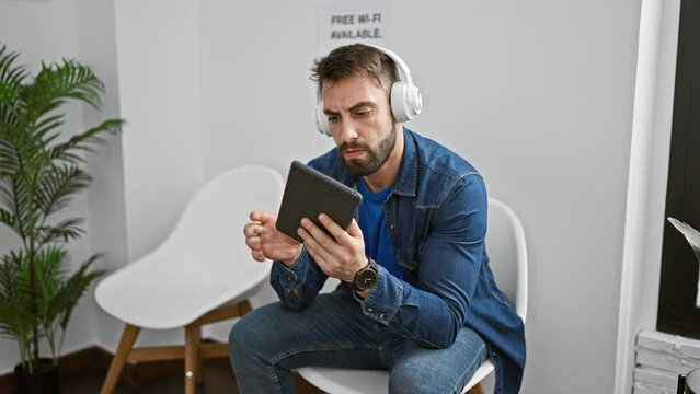 Relaxed young hispanic man enjoying tech-life in waiting room, sitting with touchpad, immersed in music through headphones