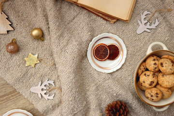Fototapeta na wymiar Bowl of cookies, cup of tea, dry oranges, pine cones, book, reading glasses and various neutral Christmas decorations on soft beige blanket. Cozy Christmas hygge. Top view.